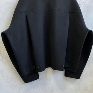 May include: A black jacket with a wide, boxy silhouette, a classic collar, and dropped shoulders. The garment features a horizontal back yoke, vertical pleats, and two small tabs at the bottom hem, each secured with a single black button.