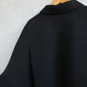 May include: A solid black jacket featuring a classic collar, a horizontal seam across the upper back, and a slightly oversized fit. The garment is displayed on a wooden hanger, showcasing the texture of the fabric and the clean, structured lines.