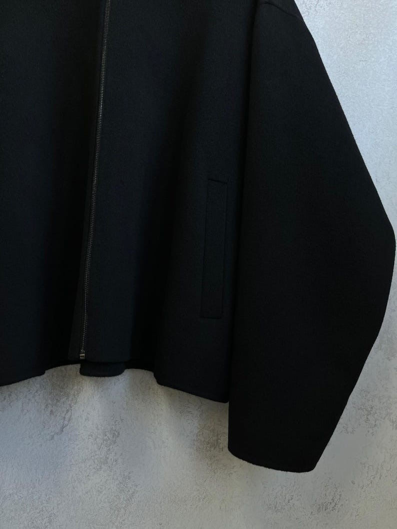 May include: A solid black jacket featuring a full-length front zipper closure, long sleeves, and a vertical slit pocket on the side. The garment has a clean, structured design with a smooth texture and a straight hemline, presented in a neutral tone.