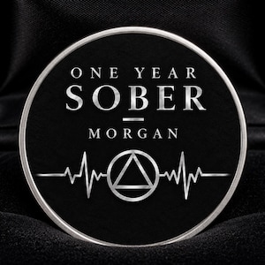 Könnte beinhalten: Eine kreisförmige schwarze Nüchternheitsmünze mit silbernem Text ONE YEAR SOBER MORGAN, einer silbernen Herzschlaglinien-Grafik und einem Dreieckssymbol in einem Kreis. Die Münze wird in einer mit schwarzem Samt ausgekleideten Box präsentiert.