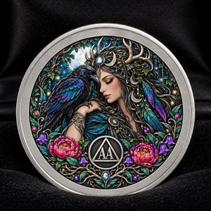 May include: A circular silver-toned medallion featuring a colourful illustration of a person with antlers and a black raven, surrounded by pink and purple flowers and foliage. A triangle symbol containing the letters AA is centred at the bottom.