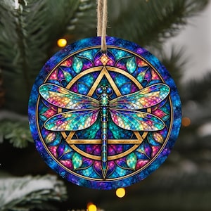 Pode incluir: Um ornamento redondo suspenso com uma ilustração vibrante de libélula com asas iridescentes. O design incorpora um padrão estilo vitral com tons de azul profundo, roxo e azul-petróleo, centrado numa moldura geométrica dourada de triângulo e círculo.