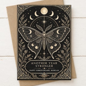 Könnte beinhalten: Eine rechteckige Grußkarte mit einer zentralen Mottenillustration, goldenen Mondphasen und Sternen auf schwarzem Hintergrund. Der Text "ANOTHER YEAR STRONGER" und "HAPPY SOBERVERSARY, MORGAN" ist in goldener Schrift gehalten und von verzierten goldenen Blumenrändern umgeben.