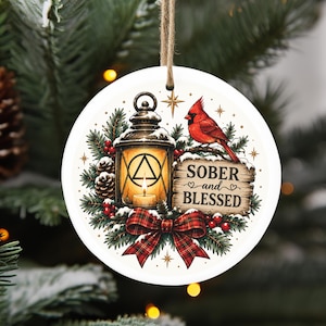 May include: A round white ceramic ornament featuring a lantern with a glowing candle and a triangle symbol, a red cardinal bird, pine branches, red berries, and a plaid bow. The text reads SOBER and BLESSED on a wooden sign background.