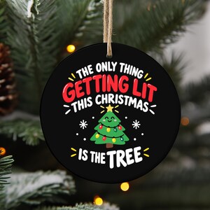 Pode incluir: Um enfeite de Natal redondo preto com o texto THE ONLY THING GETTING LIT THIS CHRISTMAS IS THE TREE em letras vermelhas e brancas. Abaixo do texto, há uma árvore de Natal verde de desenho animado com uma estrela amarela e luzes coloridas.