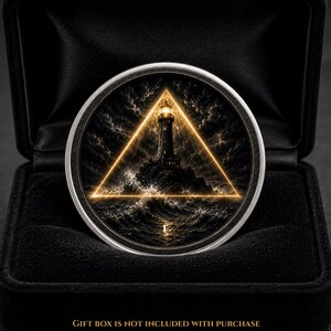 May include: A circular silver coin featuring a detailed illustration of a lighthouse on a rocky island surrounded by turbulent ocean waves, enclosed within a glowing golden triangle, presented inside a black velvet-lined display box.