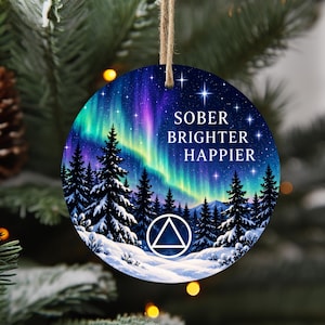 May include: A round, flat ornament featuring a vibrant aurora borealis sky over a snowy forest of pine trees. The text SOBER BRIGHTER HAPPIER is printed in white, accompanied by a white triangle symbol inside a circle, all suspended by twine.