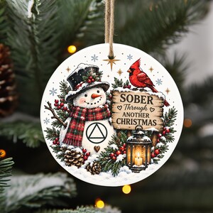 May include: A round white ceramic ornament featuring a snowman wearing a black hat and a red plaid scarf, a red cardinal bird, a lantern, and a wooden sign with the text SOBER THROUGH ANOTHER CHRISTMAS, hanging from a jute twine string.