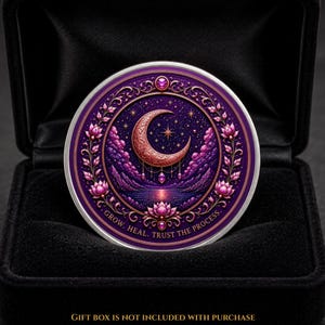 Könnte beinhalten: Eine runde Münze mit einem Halbmond- und Sternmotiv in Violett- und Goldtönen. Das Design zeigt eine Lotusblüte, Berge und den Text GROW. HEAL. TRUST THE PROCESS. Die Münze liegt in einer schwarzen Schatulle mit Samtauskleidung.