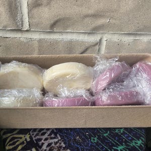May include: A rectangular tan cardboard tray containing several round bars of soap wrapped in clear plastic film. The collection includes two off-white bars and three mauve-colored bars, all arranged side-by-side within the container.