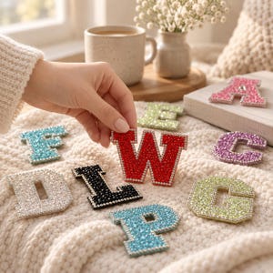 May include: A collection of alphabet letter patches covered in sparkling rhinestones in various colors including red, pink, light blue, black, white, and lime green. Each letter features a silver-toned border and is arranged on a textured fabric.