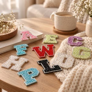 Può includere: Una collezione di toppe con lettere dell'alfabeto in stile varsity, ciascuna ricoperta di strass scintillanti. Le lettere includono A, E, C, F, W, G, H, L, D e P in vari colori come rosa, verde chiaro, viola, rosso, bianco, nero e azzurro.