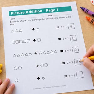 May include: A white worksheet titled Picture Addition - Page 1 with math problems involving triangles, squares, circles, and hearts. The page includes handwritten answers in boxes, alongside colorful plastic bear counters and crayons.