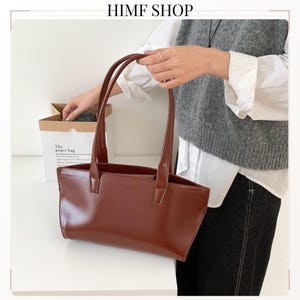 May include: A smooth, dark brown rectangular tote bag with two long, thin shoulder straps. The bag features a clean, structured design with a wide opening and a polished finish. The text HIMF SHOP is visible at the top of the image.