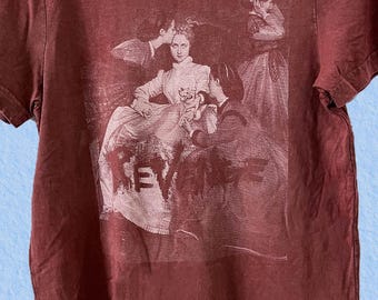 May include: A maroon-colored t-shirt featuring a faded graphic print of a vintage scene with figures and the word "REVENGE". The short-sleeved shirt has a crew neck and a slightly distressed appearance, suggesting a vintage or worn aesthetic.