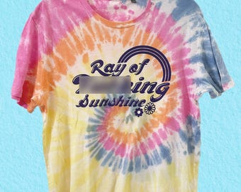 May include: A tie-dye t-shirt with a spiral pattern in shades of pink, orange, yellow, and blue. The shirt features the text "Ray of Sunshine" in a dark blue font with a matching arched design.