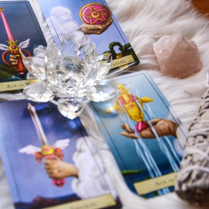 May include: A collection of tarot cards featuring illustrations of swords, pentacles, and a hand holding a scepter. A clear crystal lotus flower, a rose quartz crystal, a bundle of sage, and an amethyst crystal are also present.