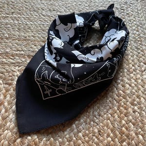 Puede incluir: Bandana negra con un dise&ntilde;o blanco de gatos y nubes. La bandana tiene un borde blanco y un s&iacute;mbolo blanco de cuatro espadas en el centro.