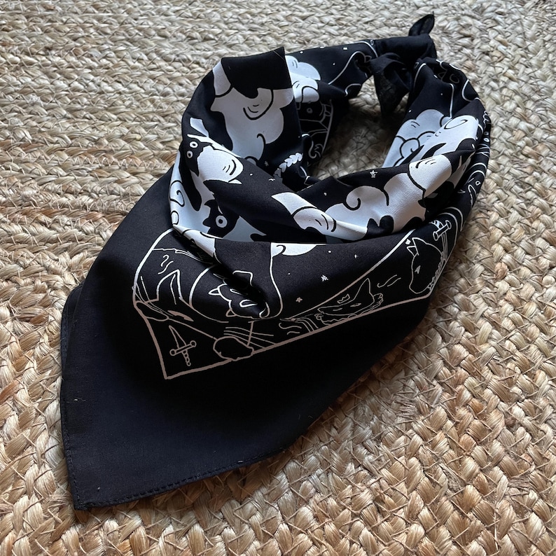 Puede incluir: Bandana negra con un dise&ntilde;o blanco de gatos y nubes. La bandana tiene un borde blanco y un s&iacute;mbolo blanco de cuatro espadas en el centro.