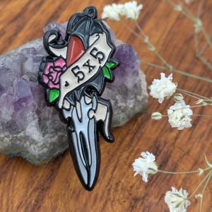 Może przedstawiać: Czarny emaliowany pin w kształcie sztyletu z bannerem z napisem "5x5". Pin ma czerwony akcent, różową różę i zielone liście. Pin jest osadzony na fioletowym kamieniu i białych kwiatach.