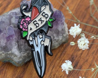 May include: A black enamel pin in the shape of a dagger with a banner that reads "5x5". The pin features a red accent, a pink rose, and green leaves. The pin is set against a purple stone and white flowers.