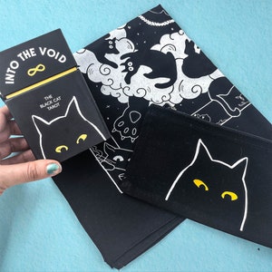 Puede incluir: Caja de baraja de tarot en blanco y negro con el texto "Into the Void" y "The Black Cat Tarot". La caja est&aacute; encima de una tela blanca y negra con un dise&ntilde;o de gato.