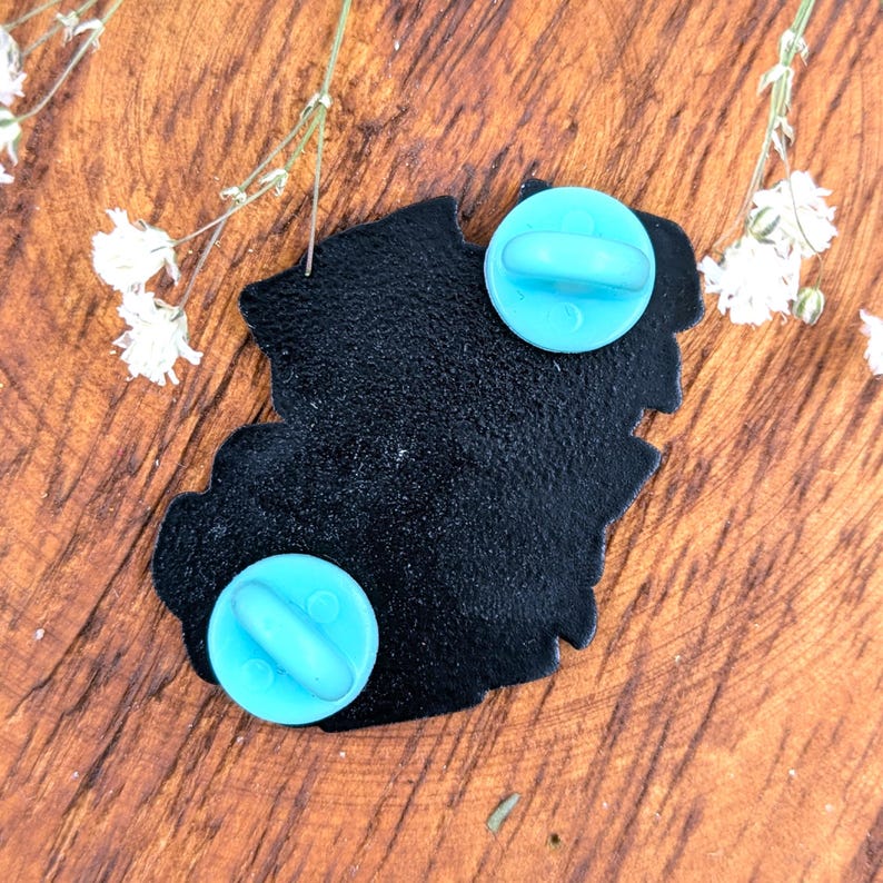 May include: Close-up of the back of a black enamel pin with two light blue pin backs. The pin is on a wooden surface with small white flowers and sprigs of dried grass.