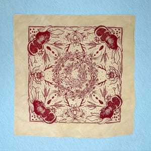 May include: A beige bandana with a red floral design featuring pomegranates and other botanical elements. The design is symmetrical and centered on the bandana.