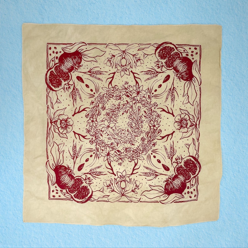 May include: A beige bandana with a red floral design featuring pomegranates and other botanical elements. The design is symmetrical and centered on the bandana.