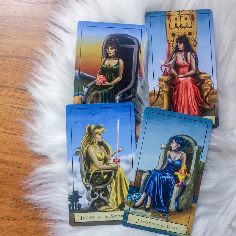 May include: Four tarot cards featuring illustrations of women in gowns, seated on ornate thrones. The cards are framed in blue, with text at the bottom. The cards are arranged on a white, fluffy surface, with a wooden surface visible in the background.