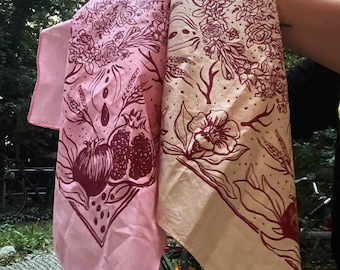 May include: Two pink and cream cotton bandanas with a floral and pomegranate design printed in maroon. The bandanas are folded and draped over a person's arm.