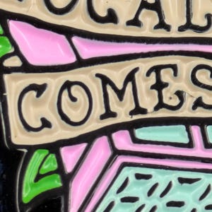 May include: Close-up of a colorful enamel pin with the word "COMES" in black letters. The pin features pink, green, and light blue geometric shapes, outlined in black, against a beige background. The text is in a bold, serif font.