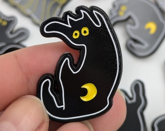 May include: Black cat-shaped enamel pin with a white outline, yellow eyes, and a yellow crescent moon. The pin is held in a hand, with other similar pins visible in the background. The cat has a whimsical, cartoonish design.