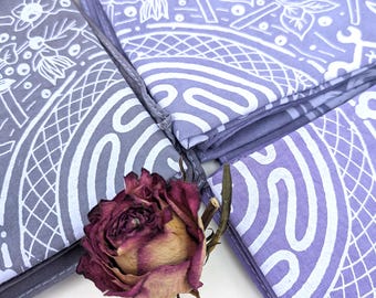 May include: Four folded lavender fabric napkins with white patterned designs, featuring floral and geometric elements. A dried, deep red rose is placed near the napkins. The napkins are stacked, showcasing the intricate patterns.