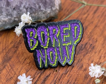 May include: A black enamel pin with the words "BORED NOW" in purple glitter lettering with a neon green outline. The pin is set against a wooden surface with white flowers and a purple stone.