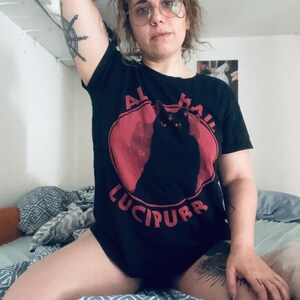 Könnte beinhalten: Ein schwarzes T-Shirt mit einem roten Kreis-Motiv, das eine schwarze Katze mit einem Kreuz auf der Stirn zeigt. Der Text "All Hail Lucipurr" ist unter dem Motiv aufgedruckt.
