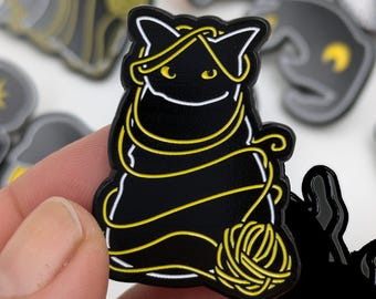 May include: A black cat enamel pin with yellow yarn wrapped around its body and head. The cat is sitting with a ball of yarn at its feet. The pin has a white outline and is set against a blurred background of similar pins.