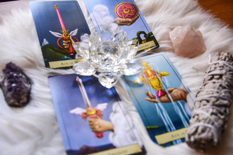 May include: A collection of tarot cards featuring illustrations of swords, pentacles, and a hand holding a scepter. A clear crystal lotus flower, a rose quartz crystal, a bundle of sage, and an amethyst crystal are also present.