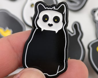 May include: A black and white cat-shaped enamel pin with yellow eyes. The pin has a white face and a black body, with a white outline. Other similar pins are in the background.