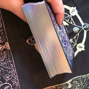 May include: A deck of tarot cards with silver edges, held in a hand. The card backs have a purple and white ornate design. The deck is on a black cloth with gold-colored symbols.