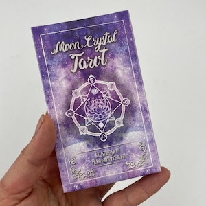 May include: A tarot card deck box with a purple galaxy design. The box features the words "Moon Crystal Tarot" in a script font, along with a circular illustration and the text "Illustrated by Alexandra Aquilina."