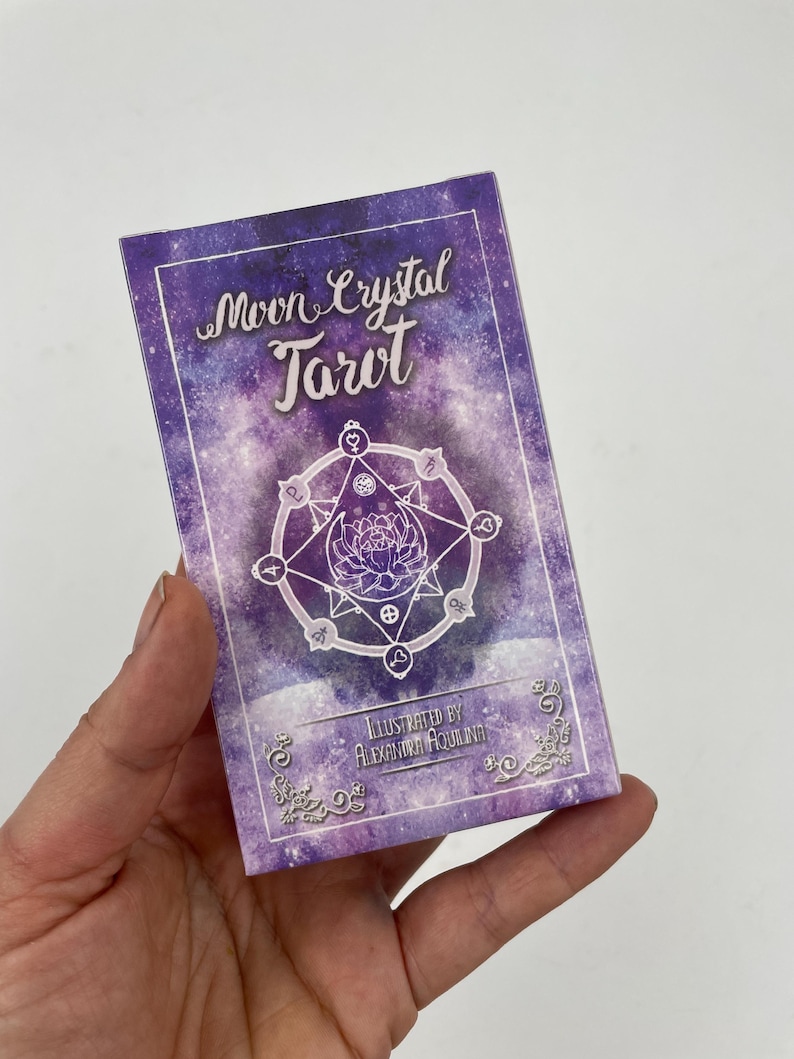 May include: A tarot card deck box with a purple galaxy design. The box features the words "Moon Crystal Tarot" in a script font, along with a circular illustration and the text "Illustrated by Alexandra Aquilina."
