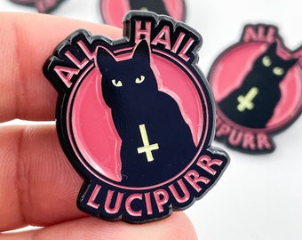 May include: A black enamel pin with a red background. The pin features a black cat with a yellow upside-down cross and the text "All Hail Lucipurr".