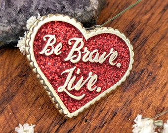 May include: Heart-shaped pin with a red glitter center and the words "Be Brave. Live." in white script. The pin has a gold-colored scalloped edge and is set against a wooden surface with small white flowers.