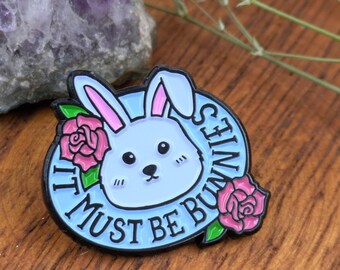 May include: Enamel pin with a light blue bunny face, pink ears, and two pink roses. The pin has the text "IT MUST BE BUNNIES" in a blue circle with a black border. The pin is on a wooden surface.