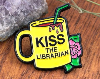 May include: A yellow enamel pin in the shape of a mug with the text "KISS THE LIBRARIAN" in black. The mug has a green straw and a pink rose with green leaves. The pin has a black border.