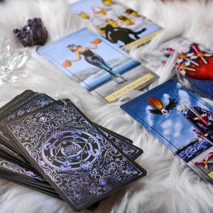 May include: A deck of tarot cards with ornate backs, alongside several face-up cards. The cards feature illustrations of figures and symbols. A crystal lotus and amethyst cluster sit on a white, fluffy surface.