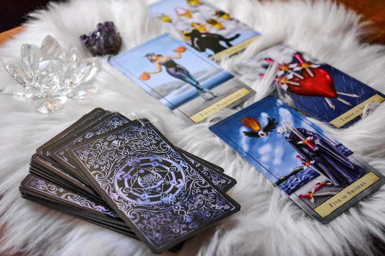 May include: A deck of tarot cards with ornate backs, alongside several face-up cards. The cards feature illustrations of figures and symbols. A crystal lotus and amethyst cluster sit on a white, fluffy surface.
