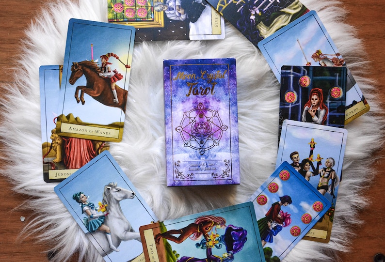 May include: A deck of "Moon Light Tarot" cards with a purple and blue celestial design. The cards are spread out on a white fluffy surface, with several cards visible, each featuring unique illustrations and titles. The deck is surrounded by other tarot cards.
