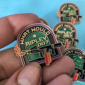 Op de afbeelding: Gouden emaille pin met een zwarte en groene achtergrond. De pin heeft een cartoon afbeelding van een pistool met een vlam en de tekst "What Would Ripley Do?" in een witte cirkel met een gouden ster.